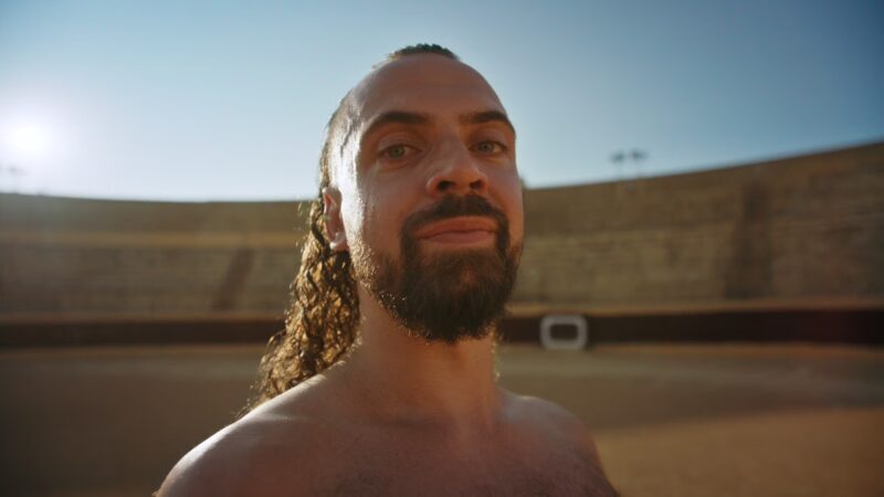een still van Giovanni die in de arena staat in Sevilla. Hij heeft geen bovenkleding aan, zijn krullende haar los hangen en een grote baard en snor. Hij kijkt van dichtbij de camera in.
