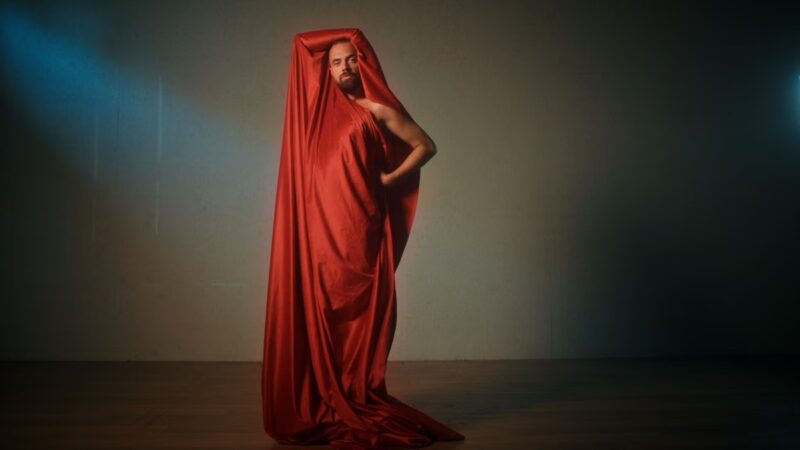 Een stil uit de documentaire. We zien Giovanni in een rood doeken gewaad geposeerd staan voor een muur.