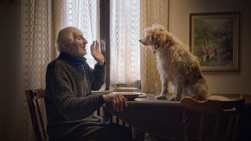 Man uit de documentaire zit aan tafel en praat tegen zijn hond die op de tafel zit.
