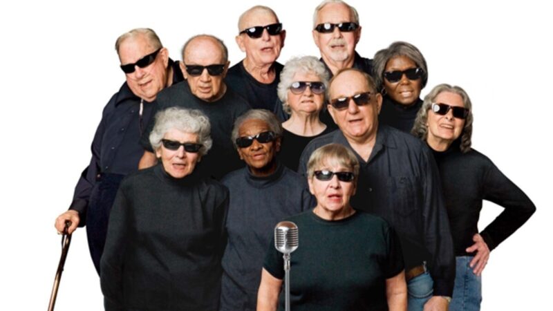 Een groep koorleden uit de documentaire zingt voor een microfoon. Ze dragen zwarte shirts en zwarte zonnebrillen.