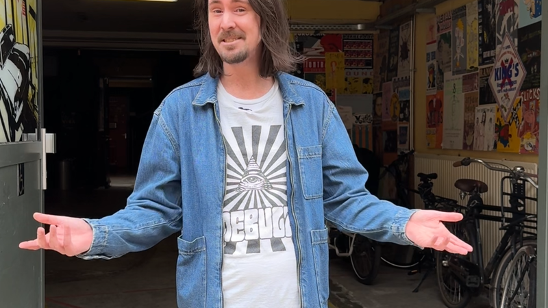 Artiestenbegeleider Mark in de deuropening van de garage van VERA. Hij heeft beide armen naar buiten gedraaid, een open houding, en kijkt vriendelijk naar de camera. Alsof hij je verwelkomd in via de artiesteningang van VERA.