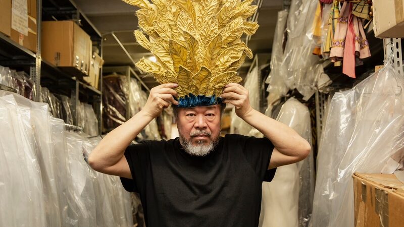 Een stil uit de documentaire waar dit artikel over gaat. we zien ai weiwei, de regisseur, met een imposante gouden tooi op zijn hoofd. Het is een wat oudere man met een grijze baard. Hij zit in een soort kledinghok, met allemaal hangers links en rechts van hem.