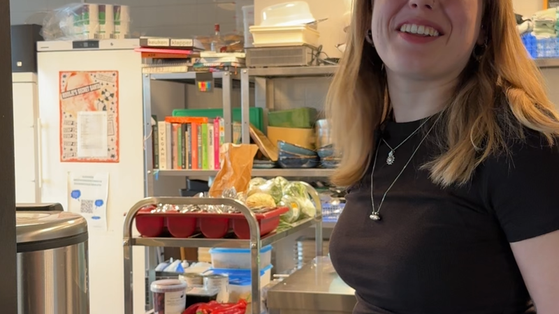 Een foto van Susanne die bij een keuken staat. Susanne is een jonge meid met los blond haar en ze kijkt lachend naar de camera. Achter haar zie je een grote professionele keuken.