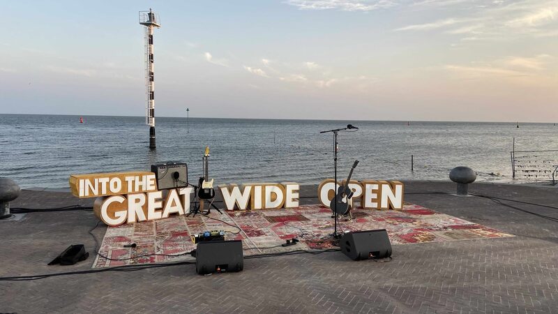 een foto van een band opstelling met op de achtergrond de zee. er staan grote letters 'into the great wide open'