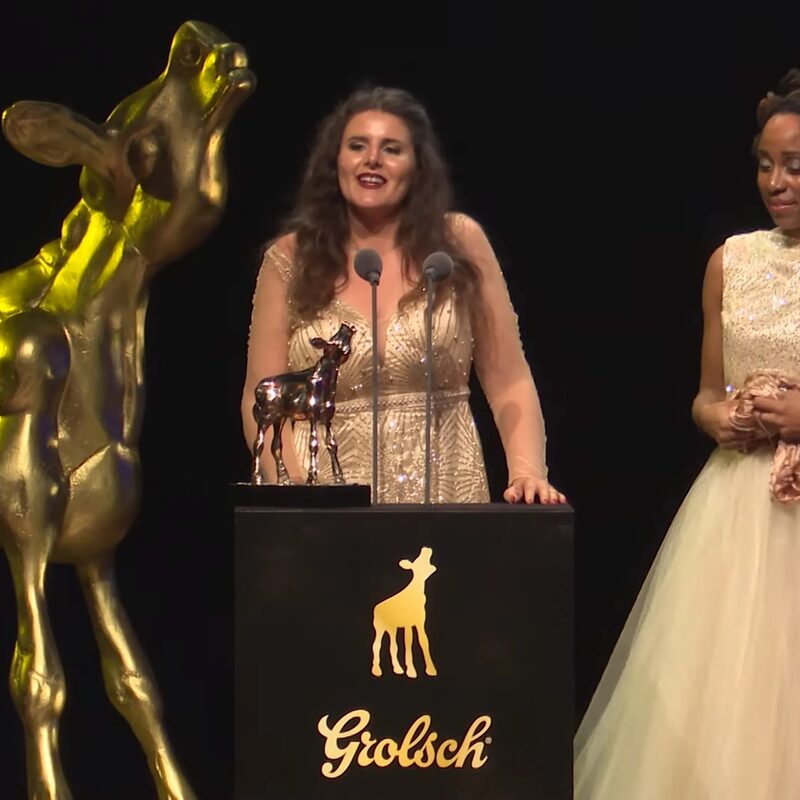 Winnares Gouden Kalf houdt een speech op het podium. Links van haar staat een gigantisch beeld van het Gouden Kalf en rechts staat een vrouw in een witte galajurk.