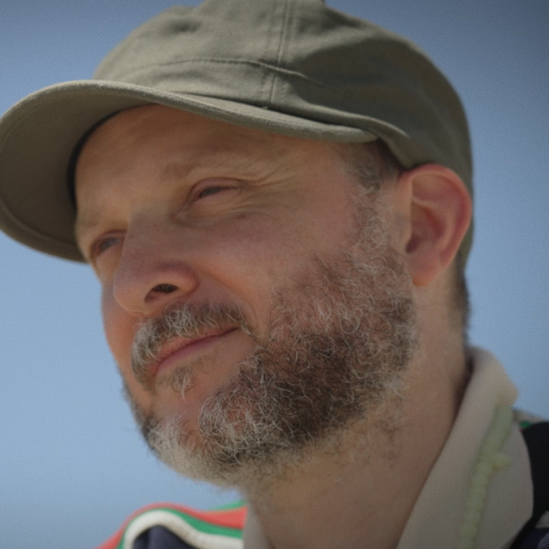 Choreograaf Sidi Larbi Cherkaoui tuurt in de verte. Hij draagt een pet en de lucht achter hem is blauw.