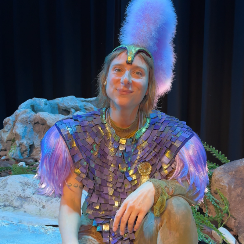 Een foto van de actrice Jip Smit, die in een paars kostuum met glitters op een soort stenen trap zit. Ze heeft een paarse verige hoofdtooi op en kijkt glimlachend naar de camera.