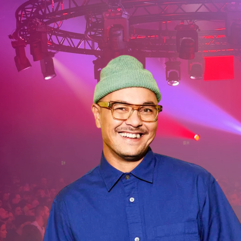 Een foto van Justin Verkijk, voor een achtergrond van een danszaal met roze licht. Justin draagt ene blauwe blouse, groene muts en bril en hij kijkt lachend de camera in.