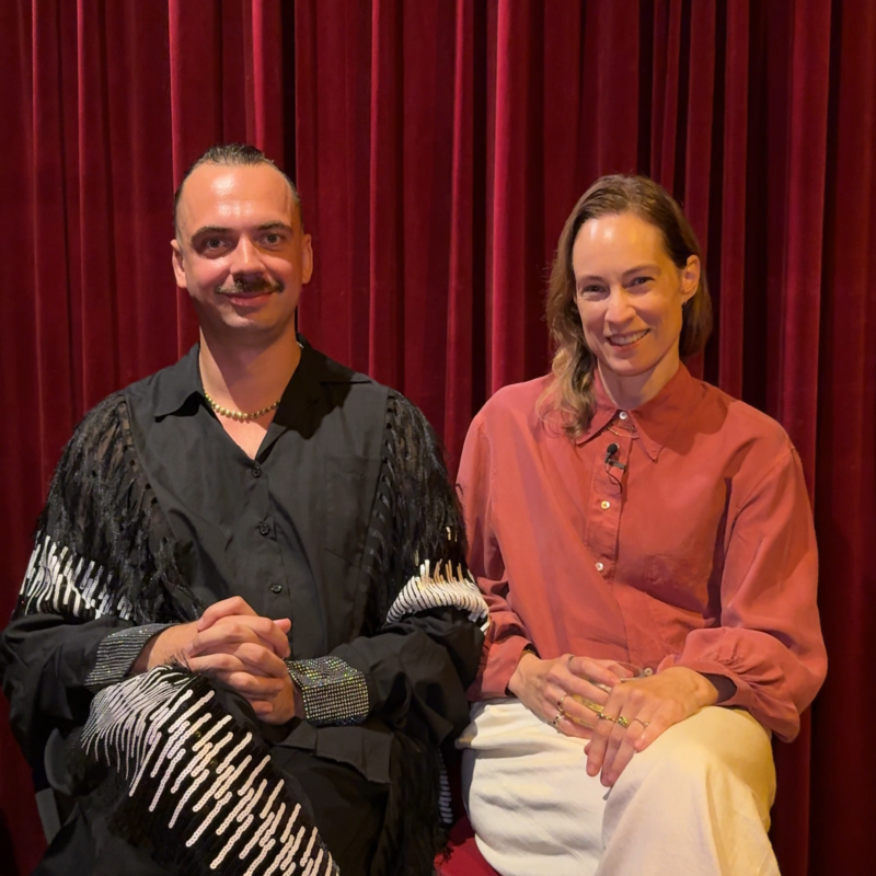 Een foto van de twee geinterviewde mensen. Ze zitten naast elkaar op een stoel voor een rood theatergordijn en kijken allebei lachend naar de camera. Giovanni zit links en heeft zwarte kleren aan, zijn haar strak naar achteren en een donkere snor. Laura zit rechts en heeft een lichtrode blouse aan, een lichte broek en blond haar wat ze los draagt.