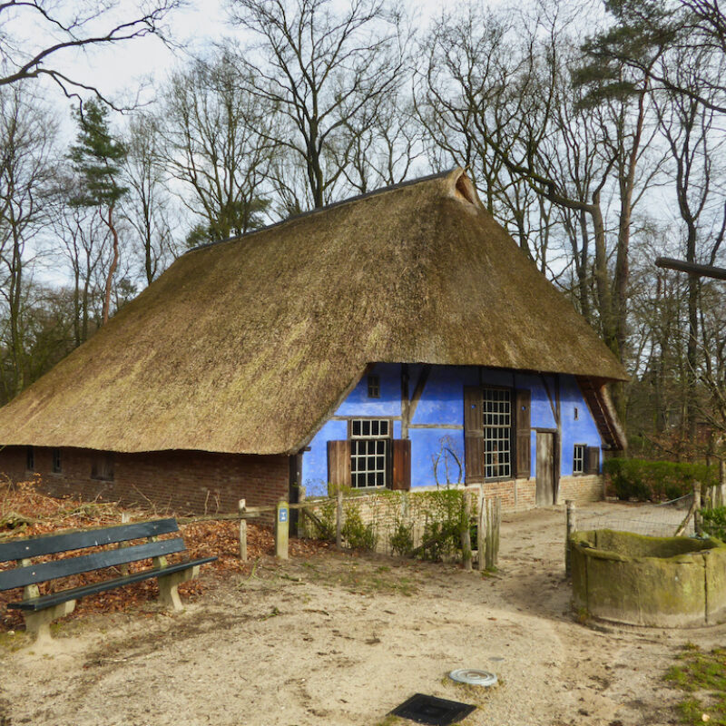 Boerderijwoning met een opvallende blauwe gevel. Naast het huis staan een waterput en twee bankjes