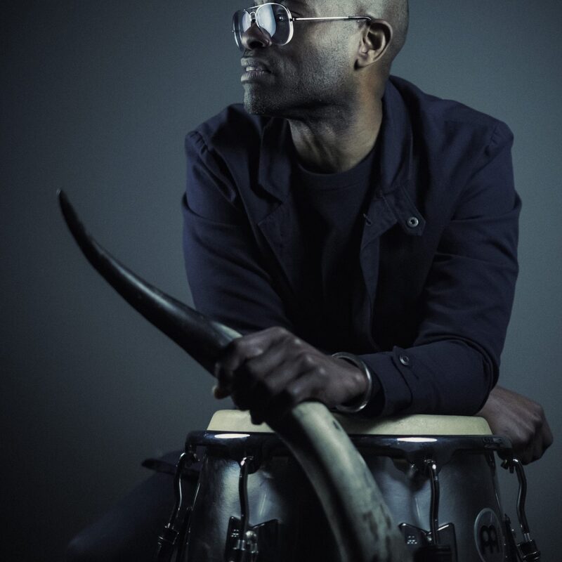 Een foto van de artiest ROel Calister. Hij heeft een spiegelende bril op en kijkt naar links. in zijn hand een soort hoorn van een dier en hij leunt op twee conga's. de foto is erg donker en hij draagt ook zwart.