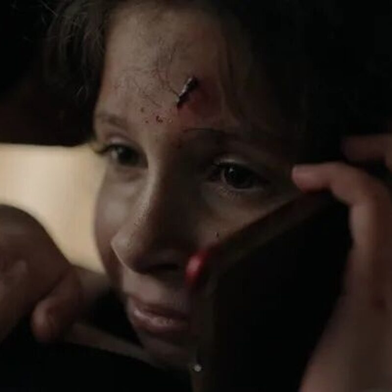 je ziet een jong meisje met een wond op haar hoofd. Ze is aan het bellen met een angstige blik.