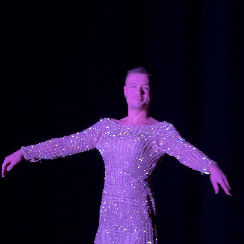 Een stilbeeld van Nick uit de voorstelling. Hij draagt een super glitterende gala jurk en kijkt trots richting de camera.