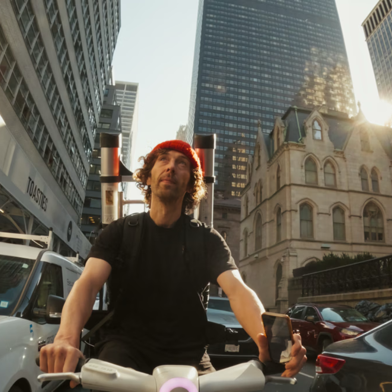 Een foto van kunstenaar street art frankey op de fiets in new york. Hij heeft zijn kenmerkende rode muts op en kijkt dromerig omhoog. Naast hem zie je druk verkeer en hele hoge gebouwen.