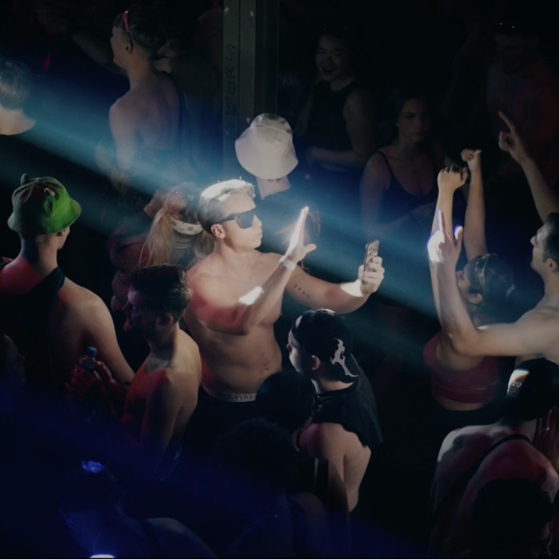 Een beeld uit de documentaire TikTok Ravers. Een jonge man met ontbloot bovenlijf staat midden op een drukke dansvloer in een lichtstraal. Hij is een andere jonge aan het filmen met een telefoon.