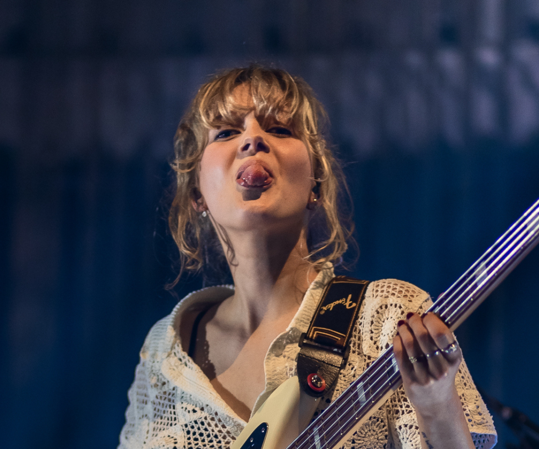 Een foto van Pieteke, een blonde meid met pony. Ze staat op een podium met een basgitaar in haar handen en steekt haar tong uit.