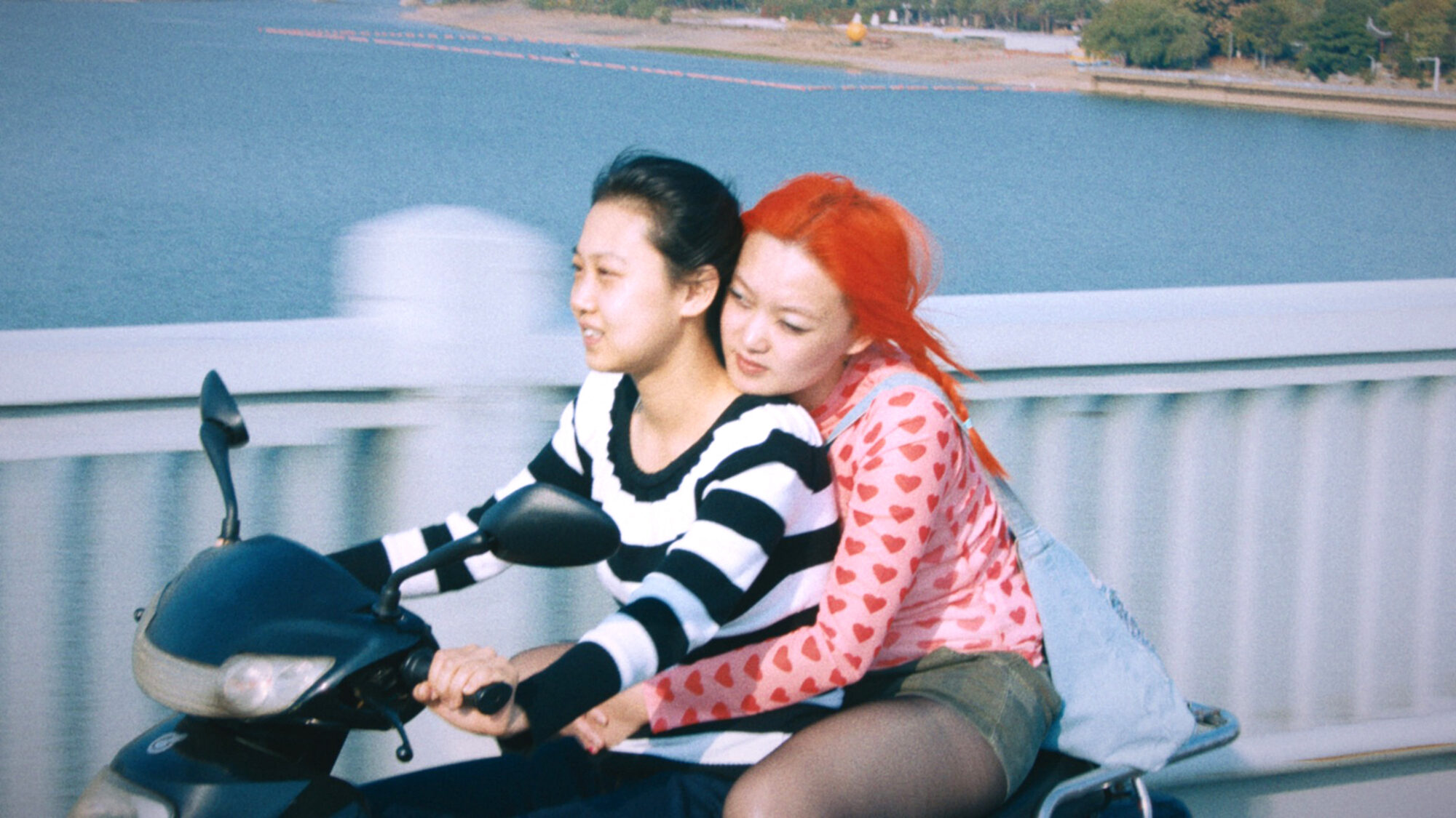 Twee Chinese actrices uit de film 1 Girl Infinite zitten samen op een scooter. Degene die rijdt, draagt een zwart/wit gestreept shirt en heeft zwart haar. Degene die achterop zit, houdt de andere persoon vast om haar middel. Ze draagt een roze shirt met rode hartjes, een groen rokje en heeft felrood haar.