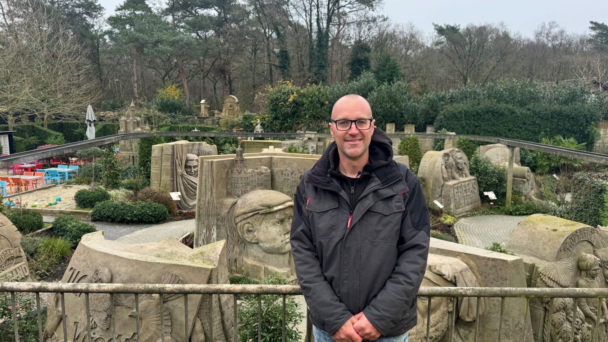 Zandkunstenaar Maxim Gazendam staat voor een railing. Achter hem zie je verschillende zandkunstwerken uit de tentoonstelling in Garderen, waaronder een groot mensenhoofd.