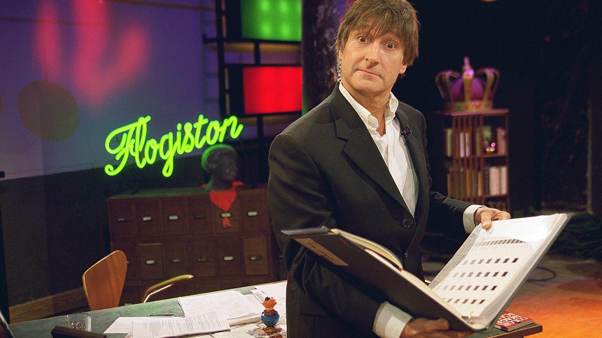 Wim T Schippers staat in een pak met een boek in zijn handen vragend kijkend naar de camera in een volle studio met een bureau en kast