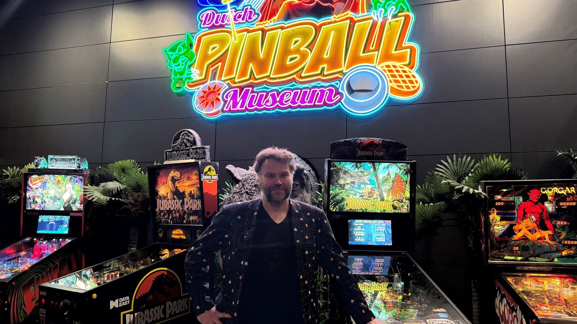 Eigenaar van het Dutch Pinball Museum, Gerard van de Sanden, staat voor vier Jurassic Park-flipperkasten en een groot neonbord in verschillende kleuren met de naam van het museum erop.