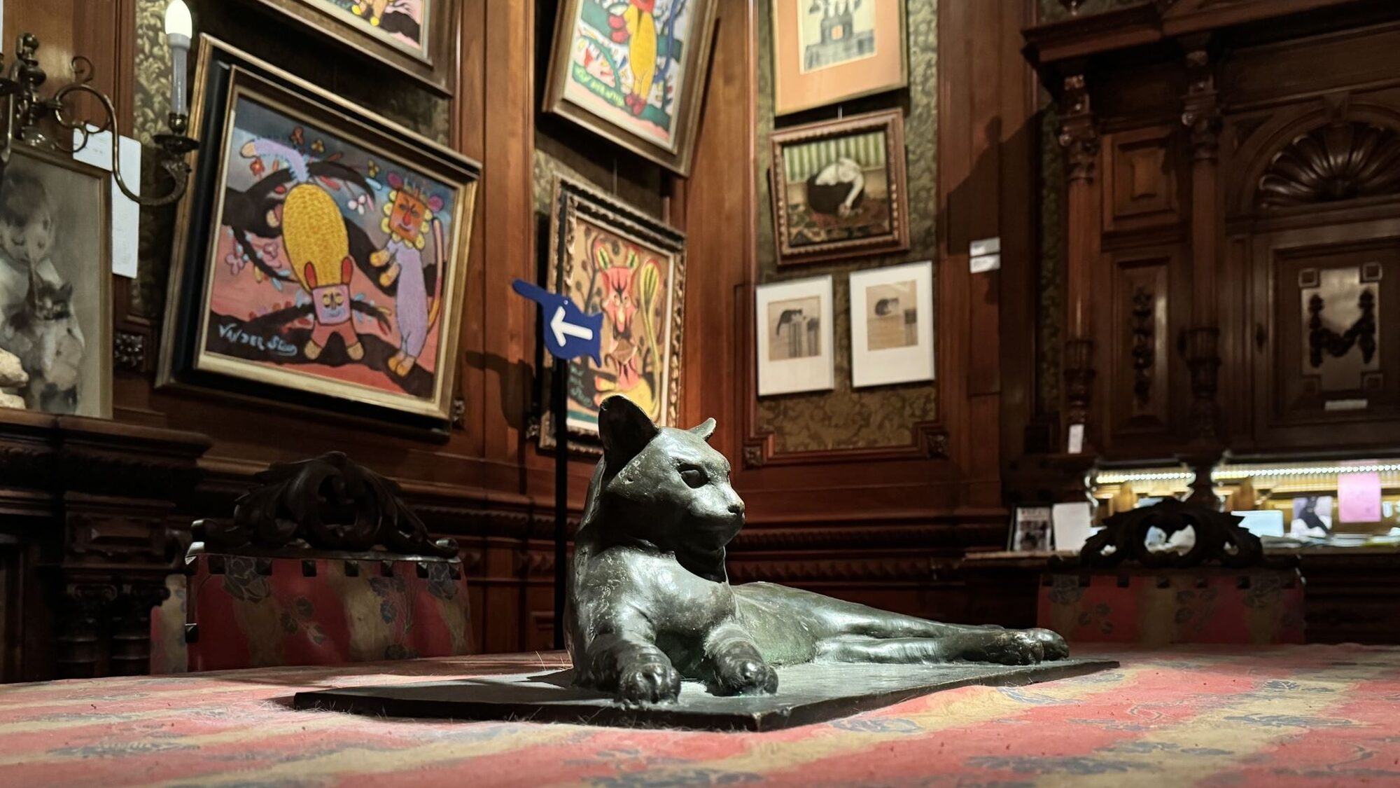 Een bronzen beeld van een liggende kat ligt op een rood beklede tafel. Op de achtergrond hangt allerlei kleurrijke schilderkunst met katten erop.