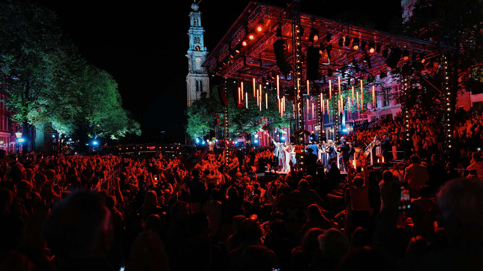 Honderden mensen staan in rood licht in het donker buiten bij de Prinsengracht in Amsterdam met een groot podium vol musici en de Westertoren op de achtergrond