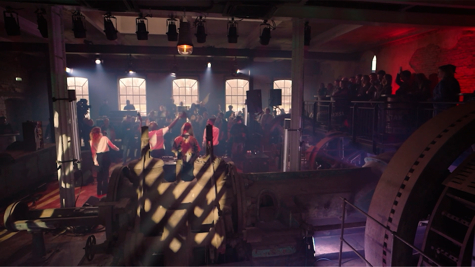 een stilbeeld van de livesessie opnames. Je ziet een grote fabriekshal waar je op de rug een band ziet spelen.