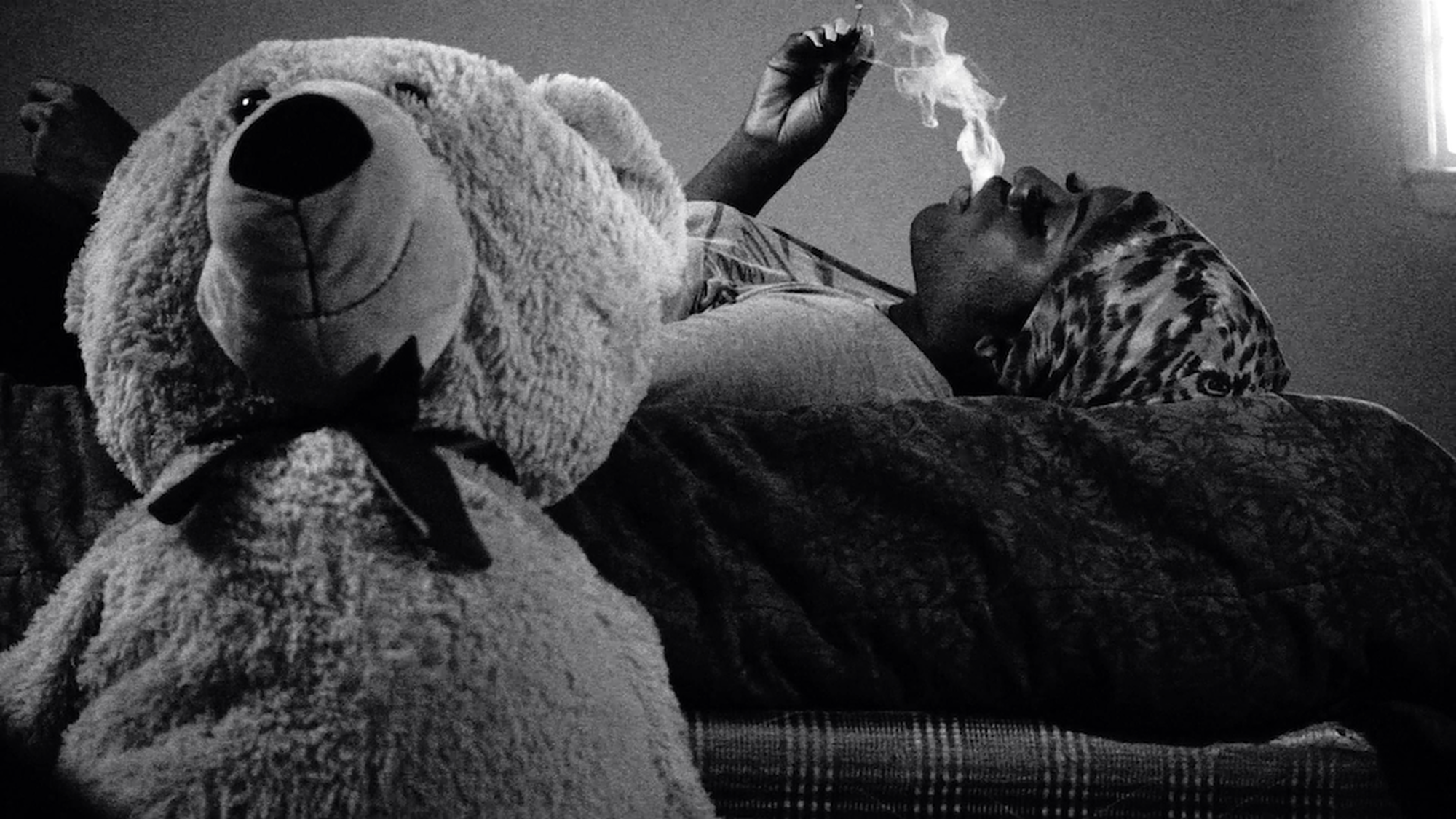 Een van de hoofdpersonen uit de documentaire ligt op bed. Ze rookt een sigaret. Naast haar staat een teddybeer.