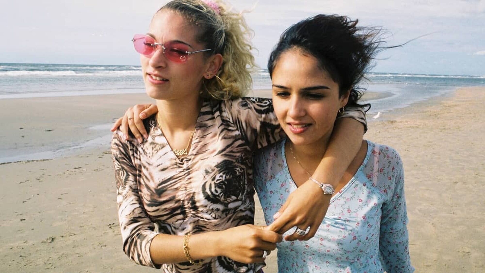Twee meiden blond en bruin lopen arm om arm lachend op het strand