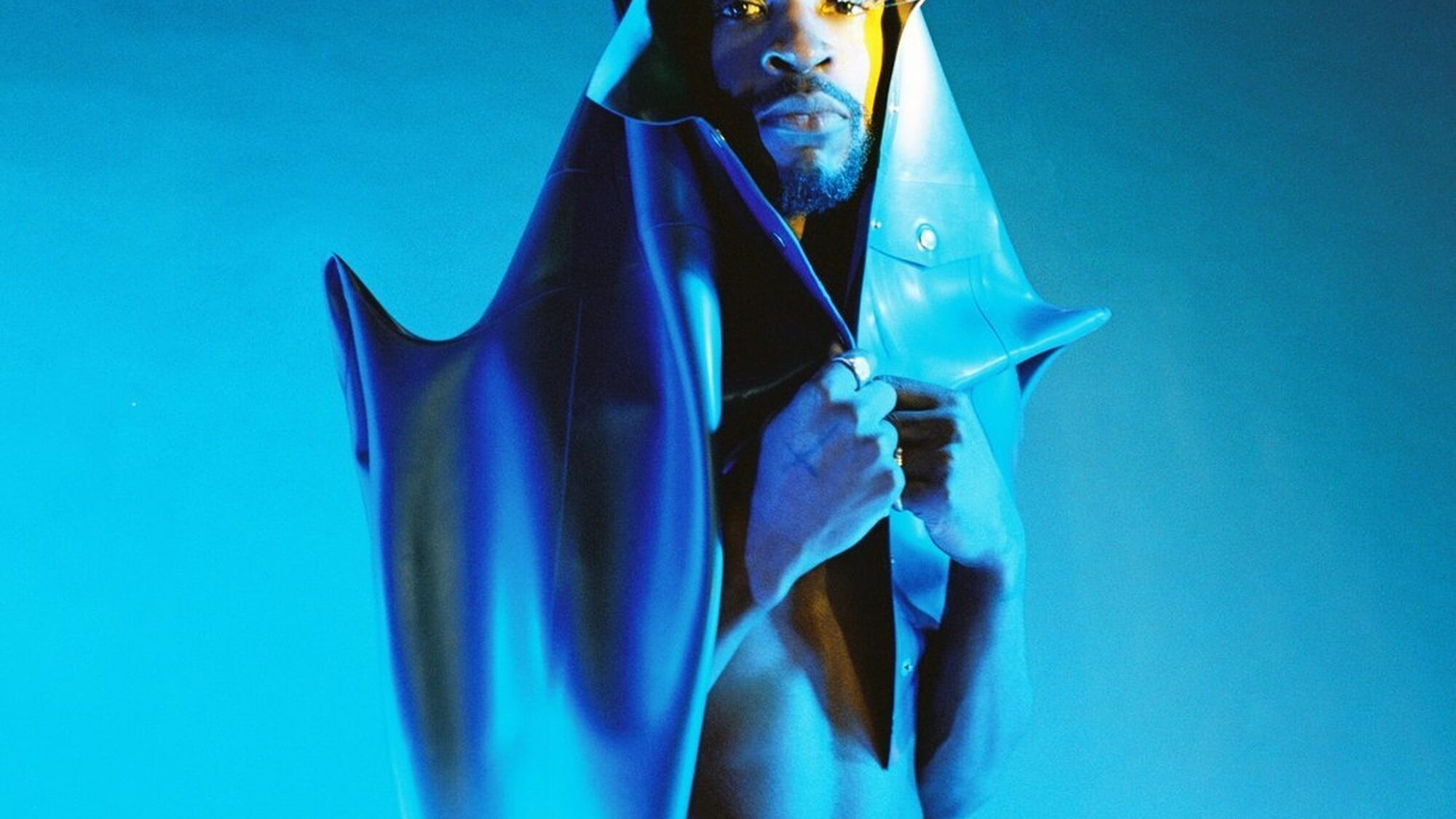 Artiest Isaï staat in blauw licht voor een blauwe achtergrond. Om zich heen draagt hij een soort sculptuur-achtige jas. Hij kijkt recht in de camera, bij zijn rechteroog draagt hij grote nepwimpers.