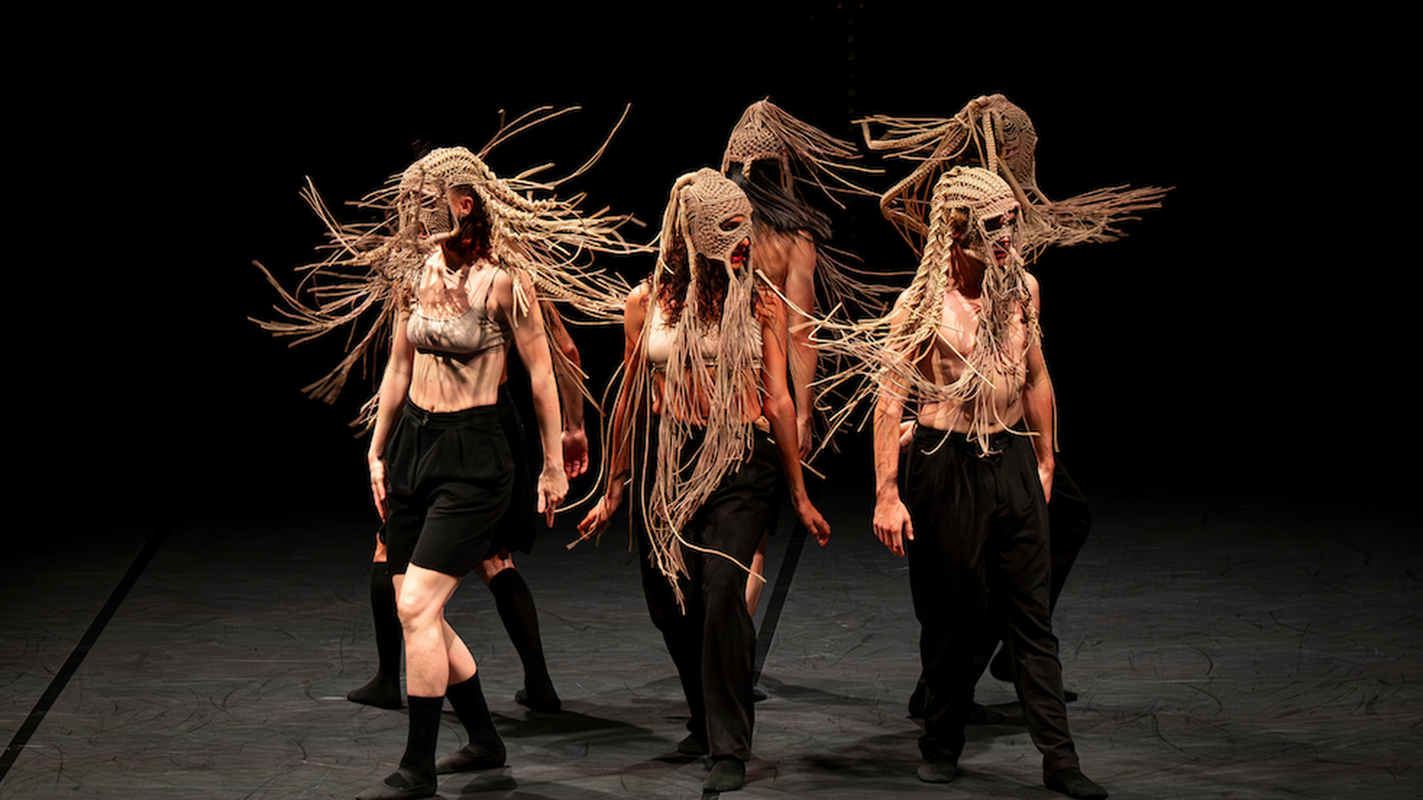 Zes dansers bewegen op het toneel. Ze dragen maskers met touwtjes die in het rond zwaaien.