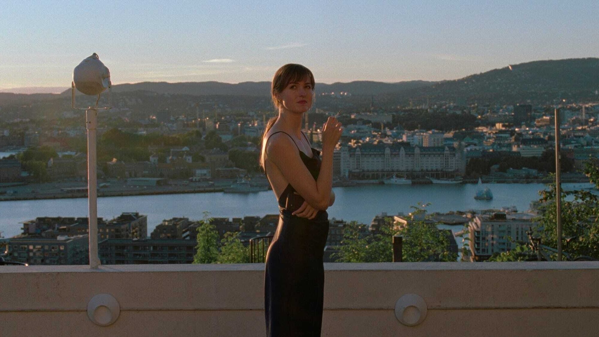 Hoofdpersonage uit de film staat in een zwarte jurk op een balkon met uitzicht over een rivier en bergen.