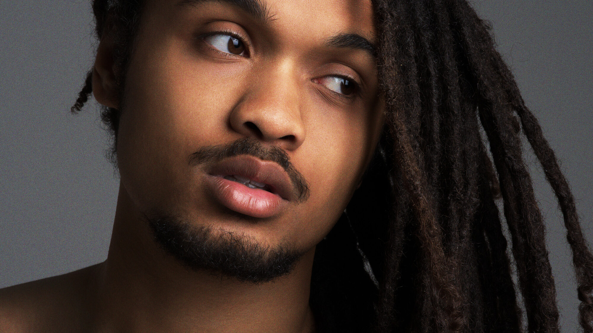 Persfoto van Jaïr Faria, een jonge jongen. De foto is van dichtbij genomen, je ziet alleen zijn hoofd tegen een grijze achtergrond. Hij heeft lang zwart haar wat naar één kant over zijn schouder hangt.