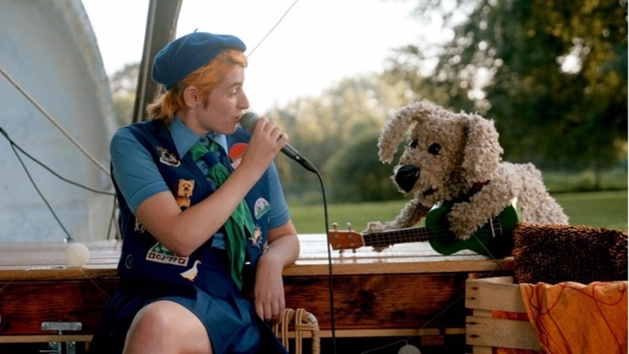 Een foto van Elmer tijdens een concert. Ze heeft een microfoon in haar hand en houdt die bij haar mond. Naast haar zit een pop van een hond met een gitaar in zijn hand. Ze zitten op een bootje.