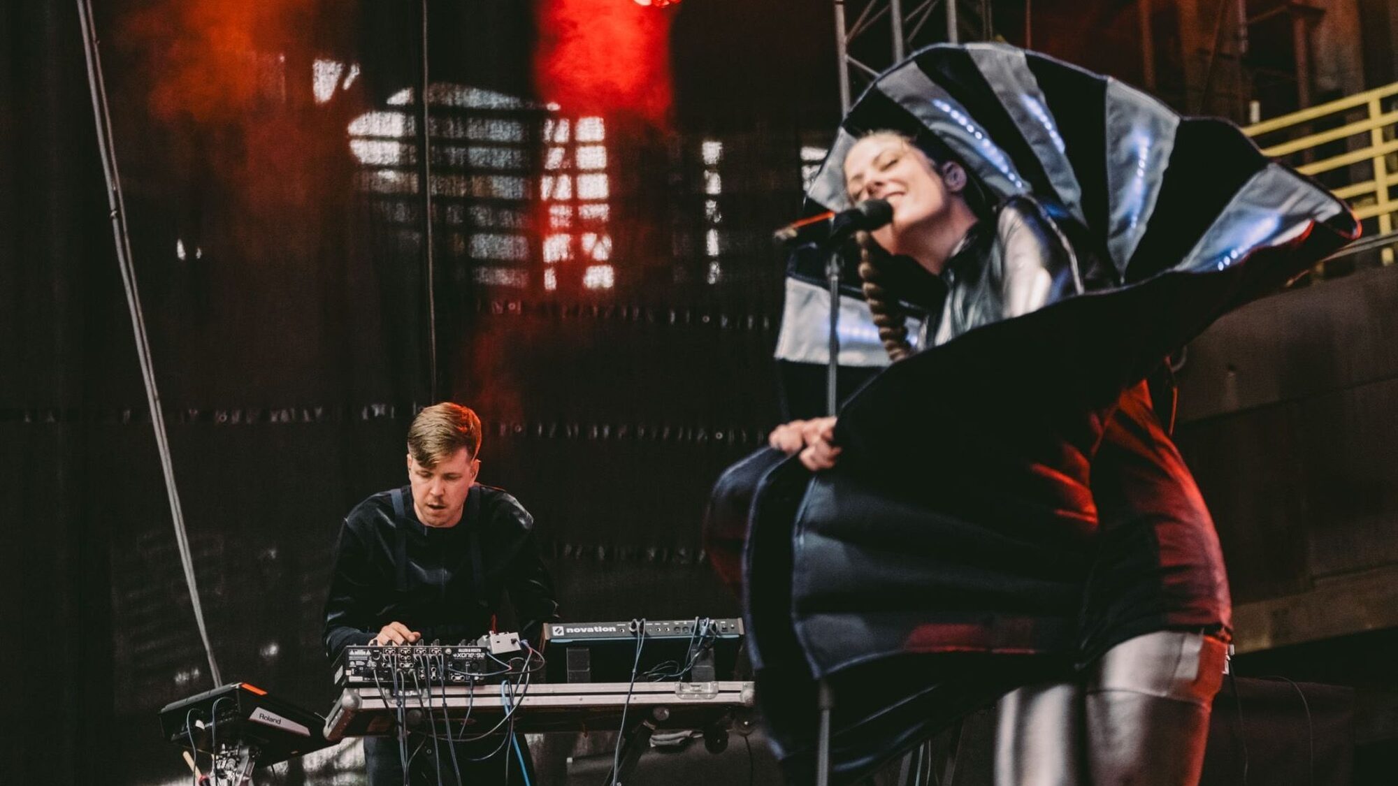 Foto van twee mensen die aan het optreden zijn. Samen vormen ze een band genaamd CUT_. Links draait een witte man achter een paneel met knoppen. Hij is in focus, heeft bruin haar en kijkt geconcentreerd. Rechts zingt een vrouw met haar ogen dicht. Ze is niet on focus en draagt een alienachtige outfit die helemaal om haar heen is gewikkeld. Het lijkt alsof ze uit een soort blad komt.