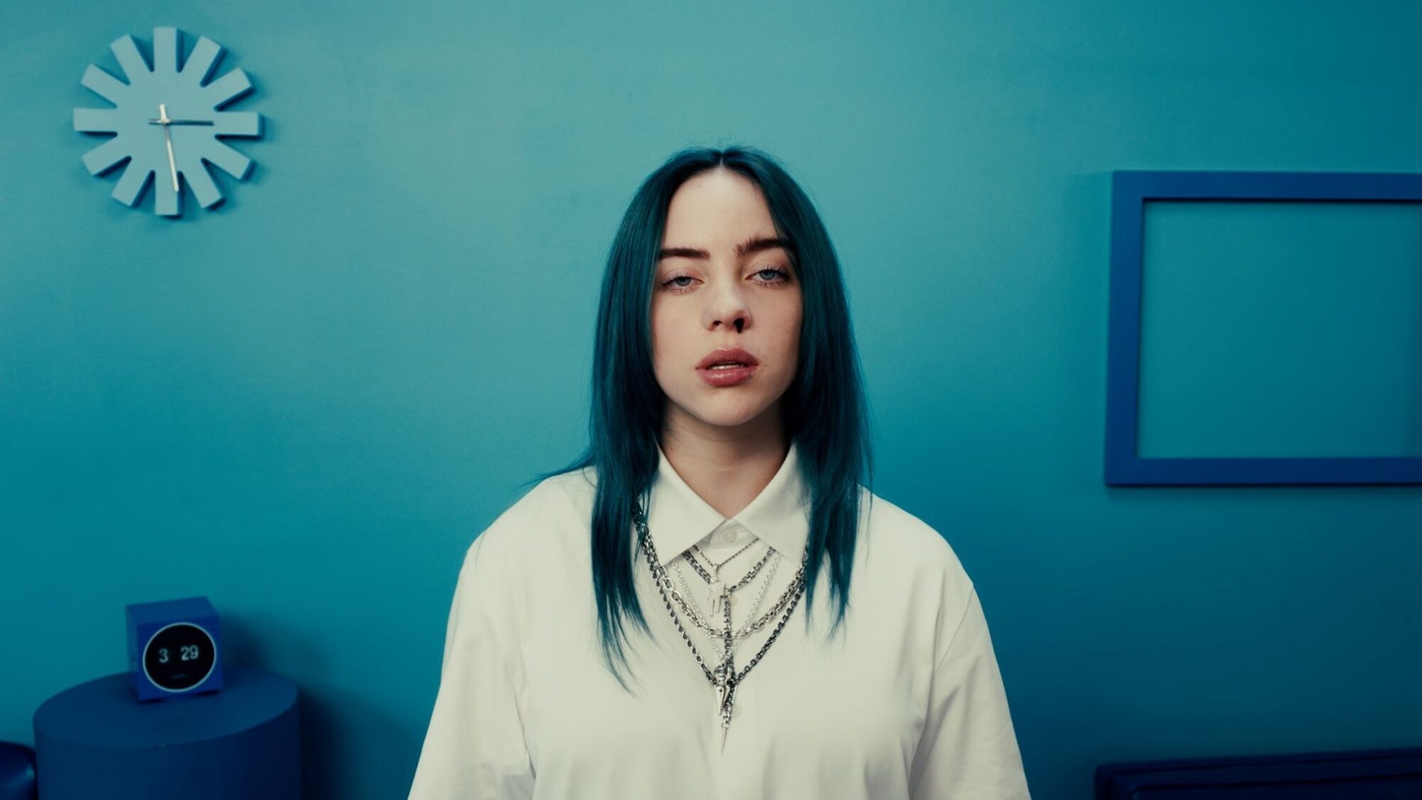 Zangeres Billie Eilish staat voor een blauwe muur. Ze heeft blauw haar, draagt een wit overhemd met kettingen erover en er komt bloed uit haar neus.