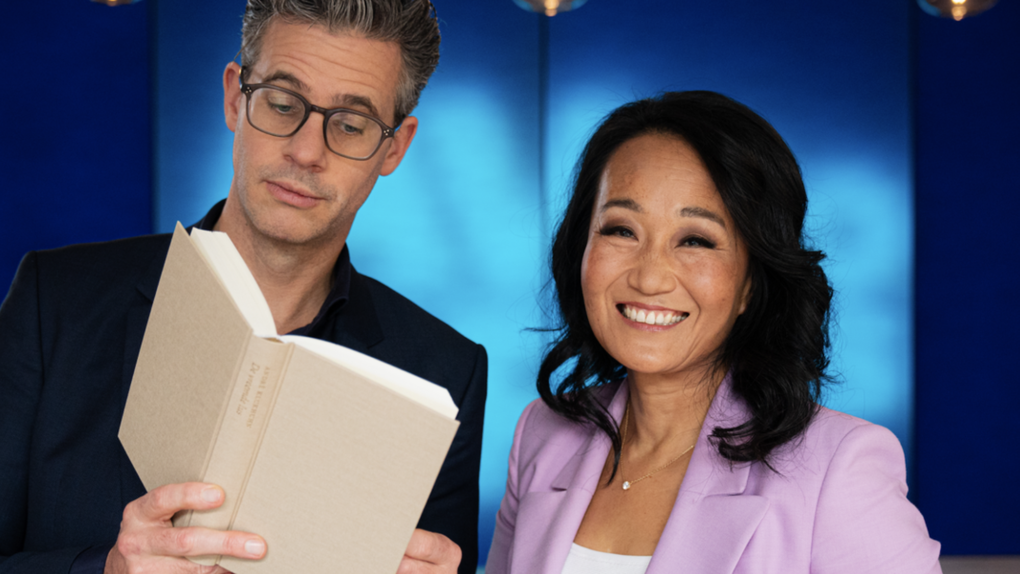 Man en vrouw staan naast elkaar en de man houd een boek vast en de vrouw lacht naar de camera met een blauwe achtergrond