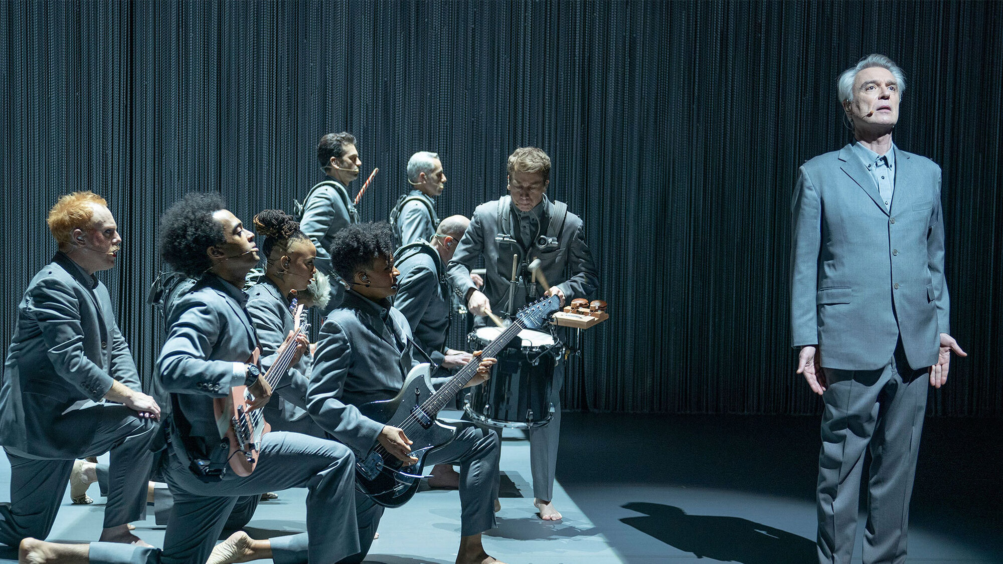 Zanger David Byrne staat op het podium in het grijs met een in grijs geklede band naast zich