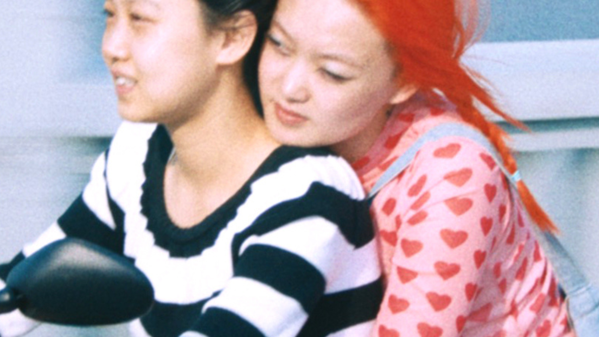 Twee Chinese actrices uit de film 1 Girl Infinite zitten samen op een scooter. Degene die rijdt, draagt een zwart/wit gestreept shirt en heeft zwart haar. Degene die achterop zit, houdt de andere persoon vast om haar middel. Ze draagt een roze shirt met rode hartjes, een groen rokje en heeft felrood haar.