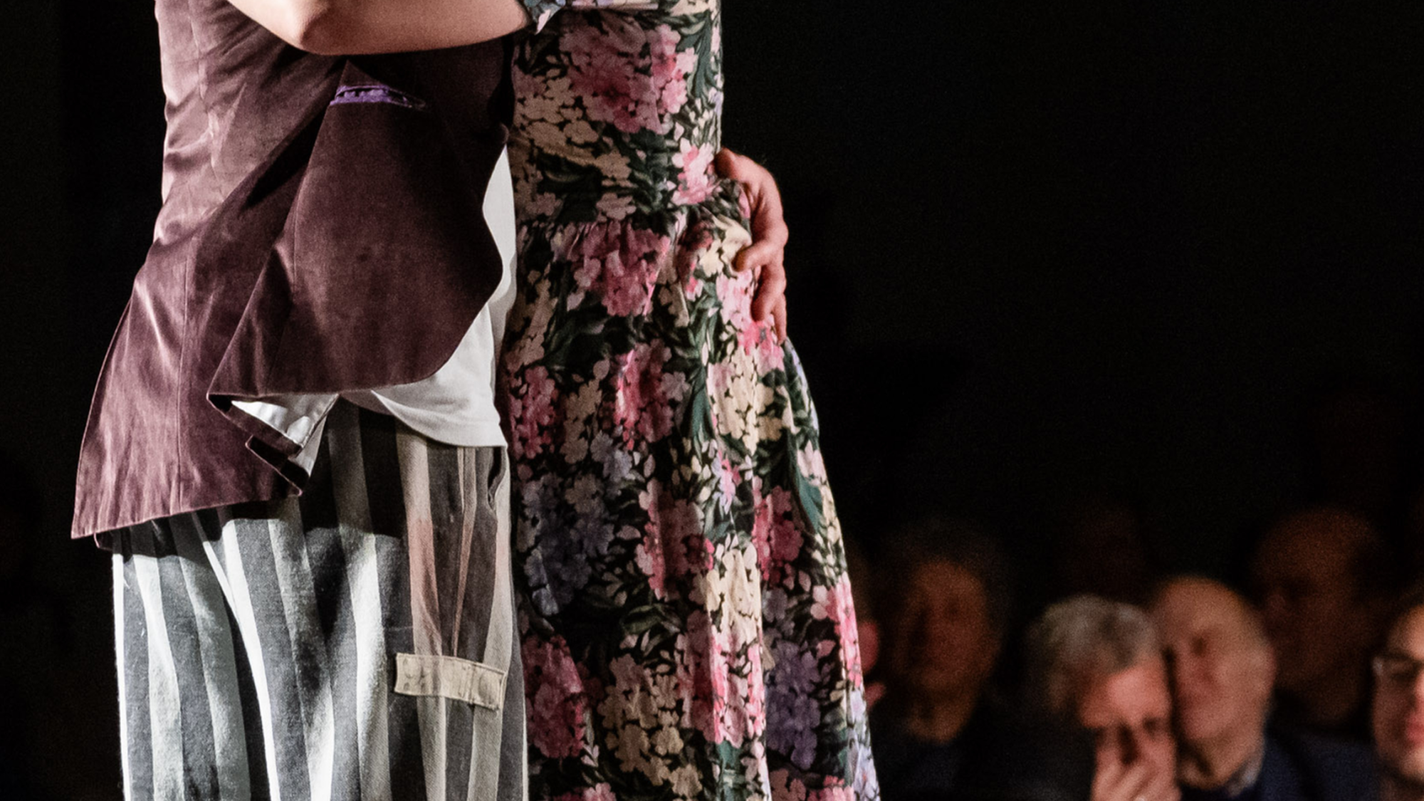 Op de foto zijn twee knuffelende personen op een podium te zien. Een persoon, de vrouw, heeft een bloemetjesjurk aan tot over haar knieën. Daaronder draait ze een loszittende grijs-gestreepte broek. De man draagt een bruin colbert. Ook hij draait dezelfde soort grijs-gestreepte broek. Naast hen op het podium staan een rode tafel en stoel.