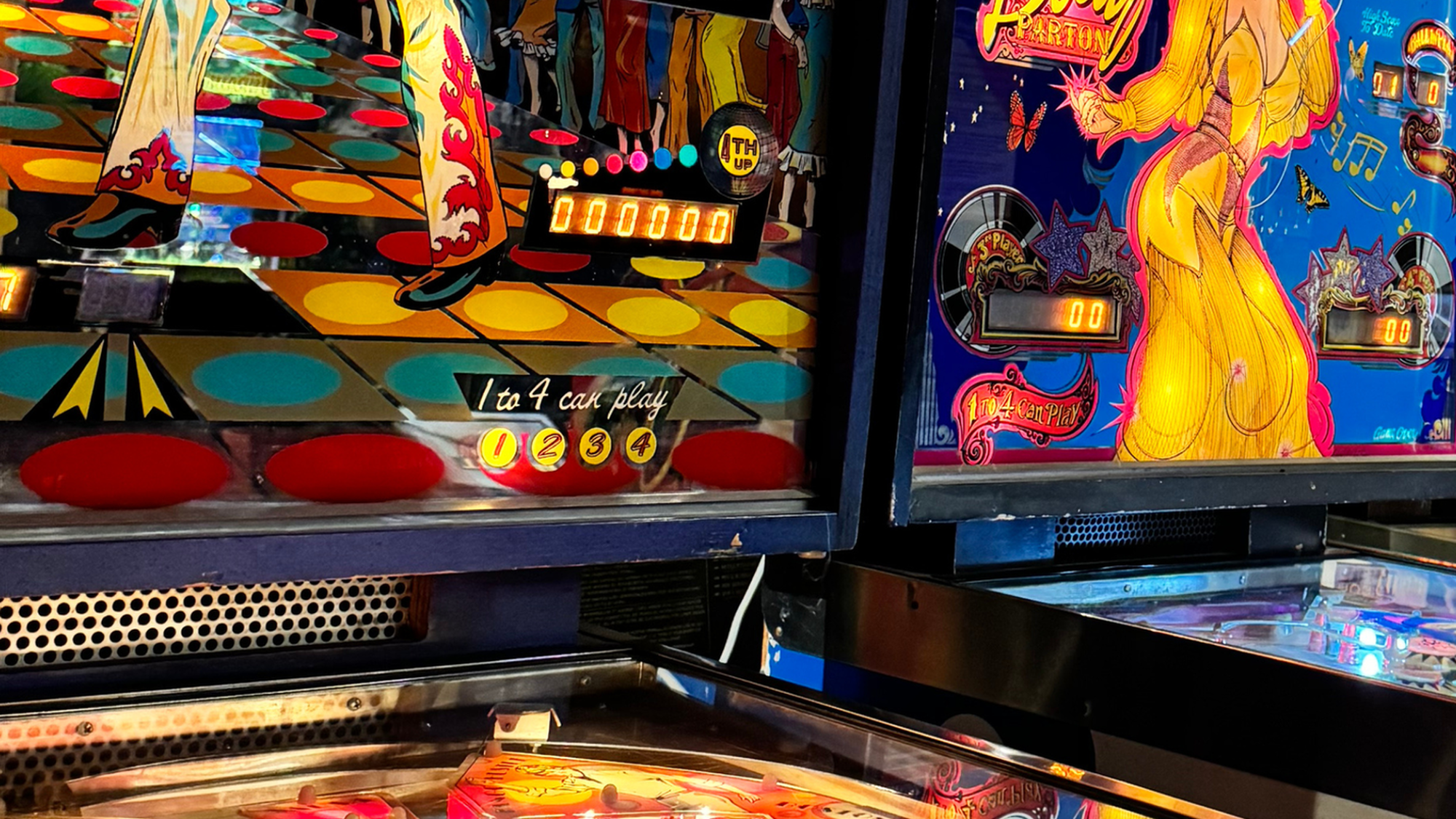 Twee lichtgevende flipperkasten naast elkaar. Links één in een discothema met een dansende man erop en rechts één met een Dolly Parton-thema.