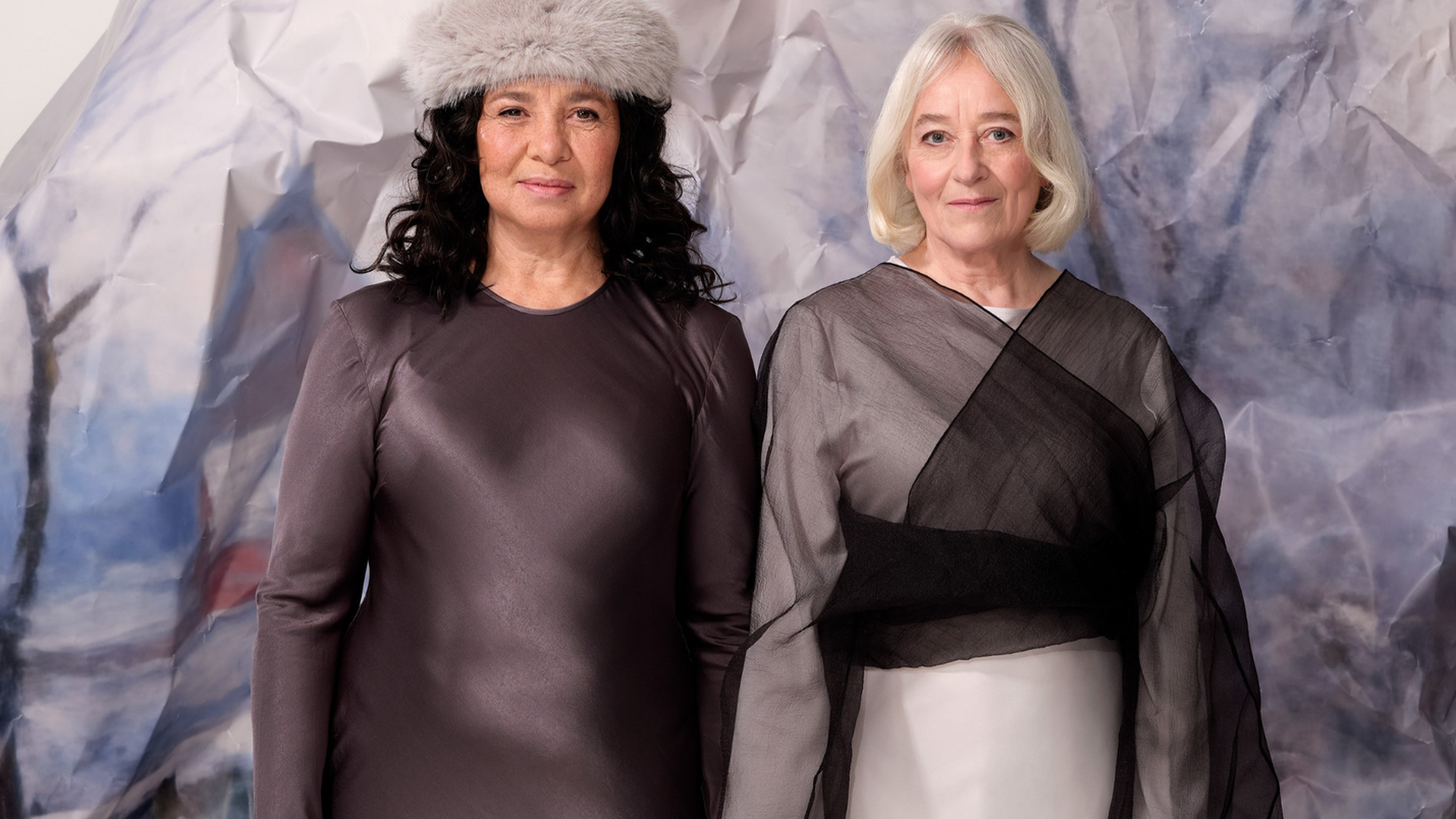 Twee vrouwen staan naast elkaar met hun vingers in elkaar verweven. Achter hen staat een ijsberg van papier. De linker vrouw heeft donkerbruin krullend haar, draagt een grijze muts en een lange bruine jurk. De rechter vrouw heeft kort, stijl en blond haar en draagt een lange zwart/witte jurk.