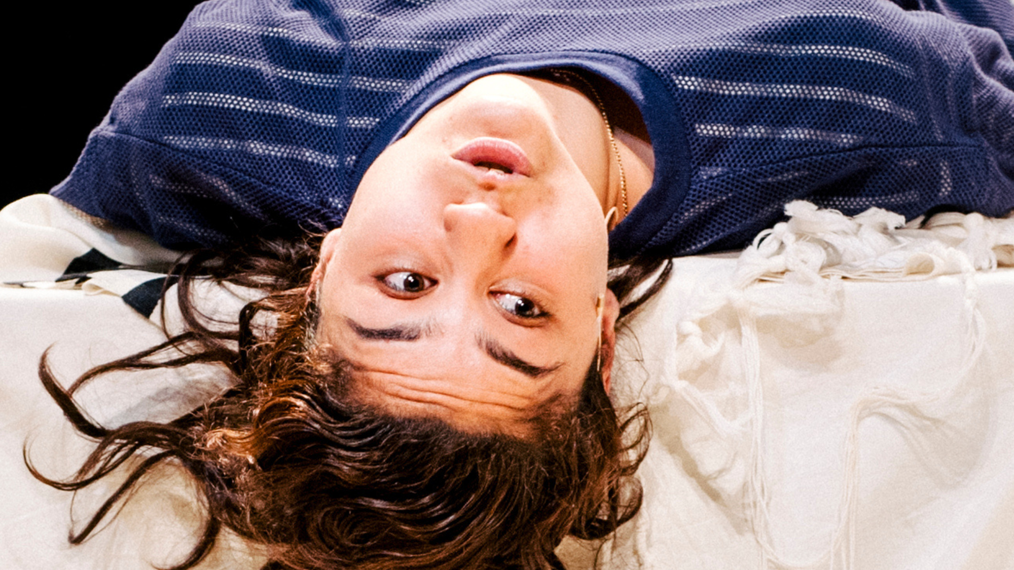 Actrice Soumaya ligt in blauwe kleding met haar rug op een tafel met een groot wit doek. Haar hoofd hangt over de tafel heen en ze kijkt naar rechts.