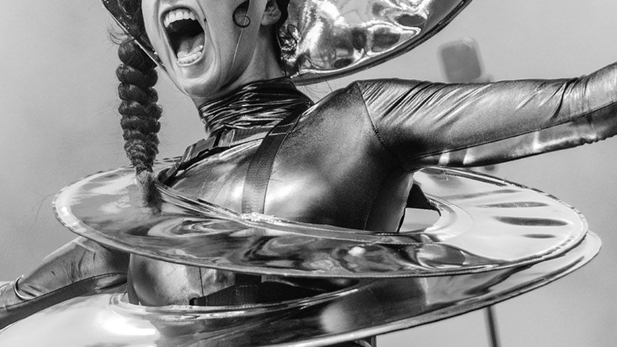 Zwartwitfoto van een vrouw die met haar armen wijd op een podium staat. Haar mond heeft ze wijd open en ze heeft een alienachtig kostuum aan dat bestaat uit een soort schotels. Op haar hoofd draagt ze ook een schotel.