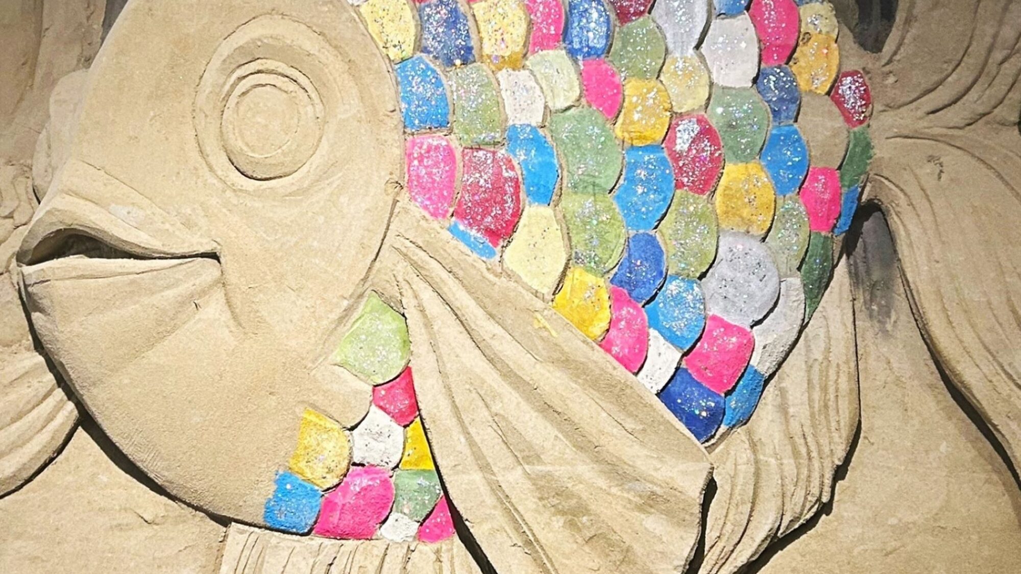 Zandsculptuur van de vis uit het kinderboek 'De mooiste vis van de zee'. Zijn kop en vinnen zijn zandkleurig (beige) en zijn schubben zijn alle kleuren van de regenboog (blauw, roze, groen, rood, wit, geel).