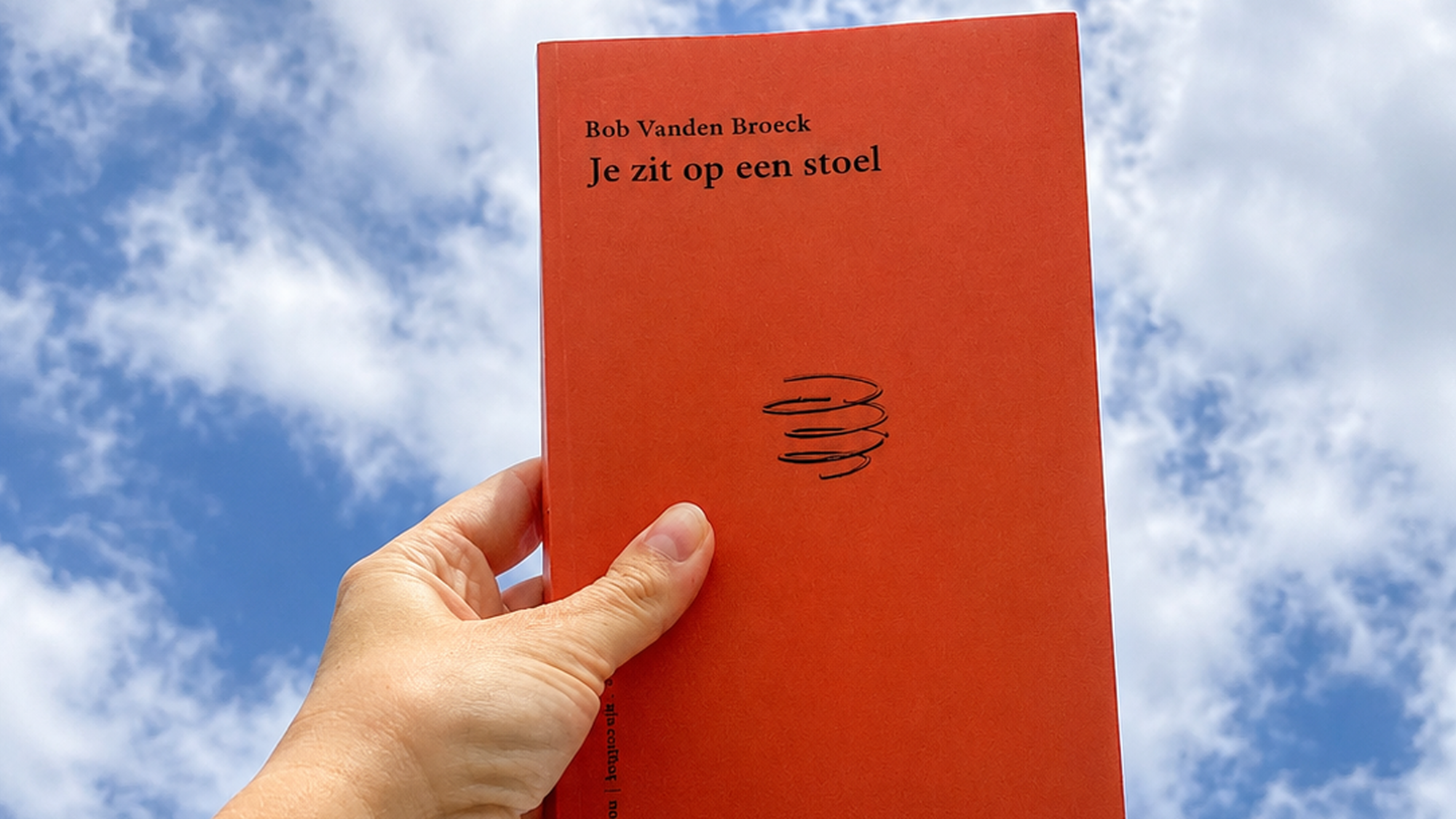Oranje boek met de titel 'je zit op een stoel' wordt vastgehouden door een witte hand met de blauwe lucht met witte wolken op de achtergrond