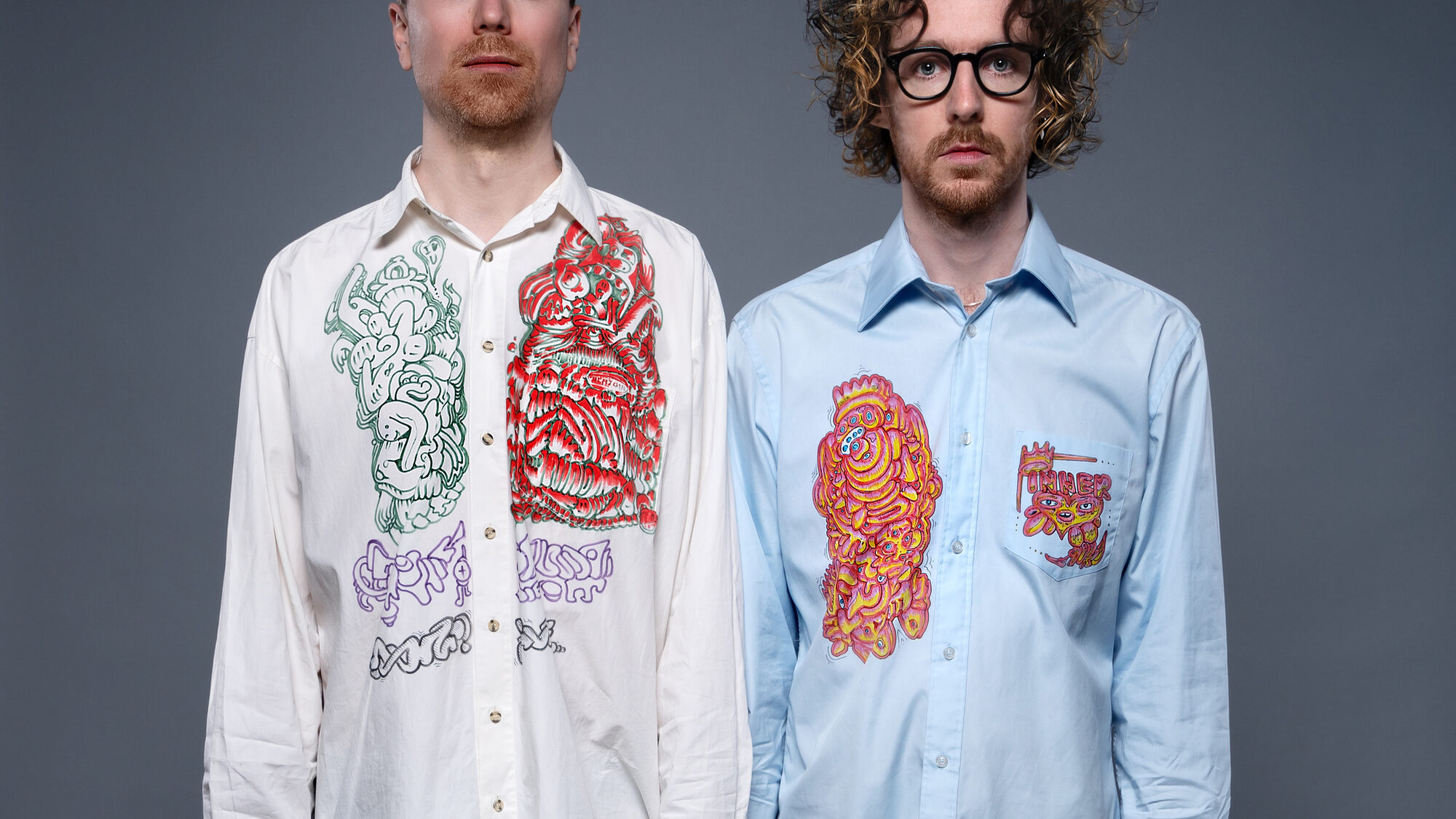 Persfoto van de band Ciao Lucifer. Er staan 2 mannen naast elkaar voor een grijze achtergrond. De linker is blond en de rechter heeft donkere krullen en een bril. Ze dragen beide een lang overhemd met drukke print erop en geen broek.