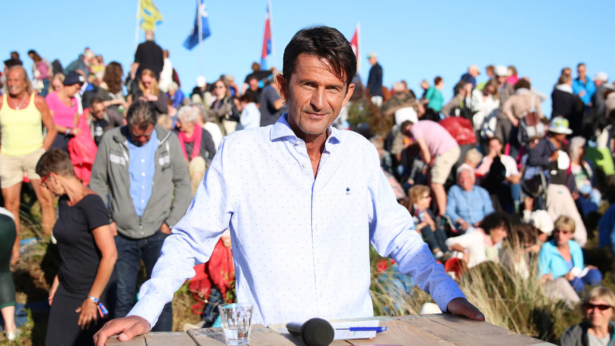 Cornald Maas staat glimlachend in een overhemd achter zijn tafel op de set van Opium op Oerol op Terschelling met heel veel mensen achter hem