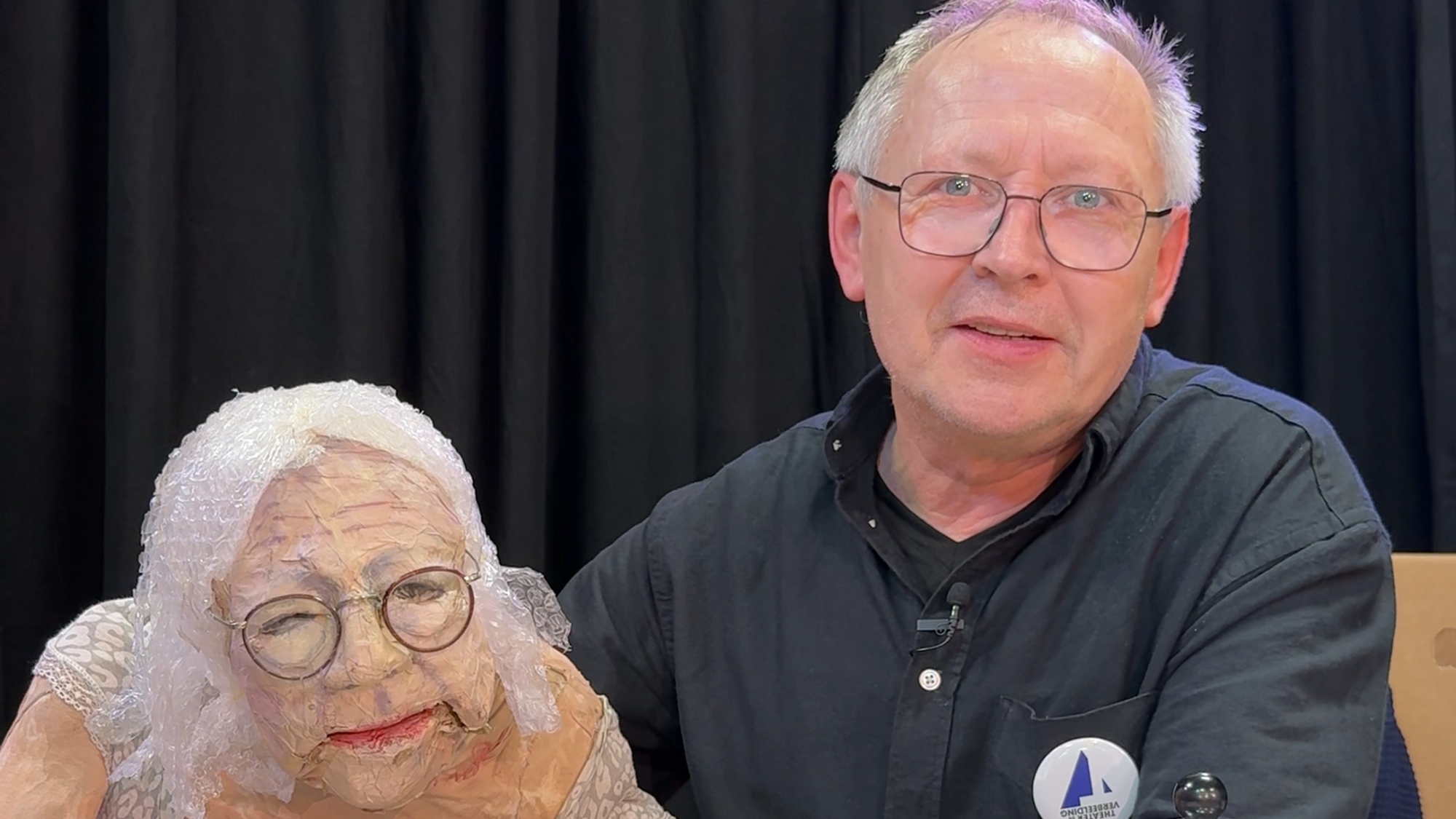 een foto van Servaes met zijn pop. Servaes zit rechts, met een zwart overhemd en een bril en grijs haar. Naast hem zit een pop van een oude vrouw, met ook een bril en bubbeltjesplastic als haar. Ze zitten achter een tafel waar ook een theepot en kopjes op staan.