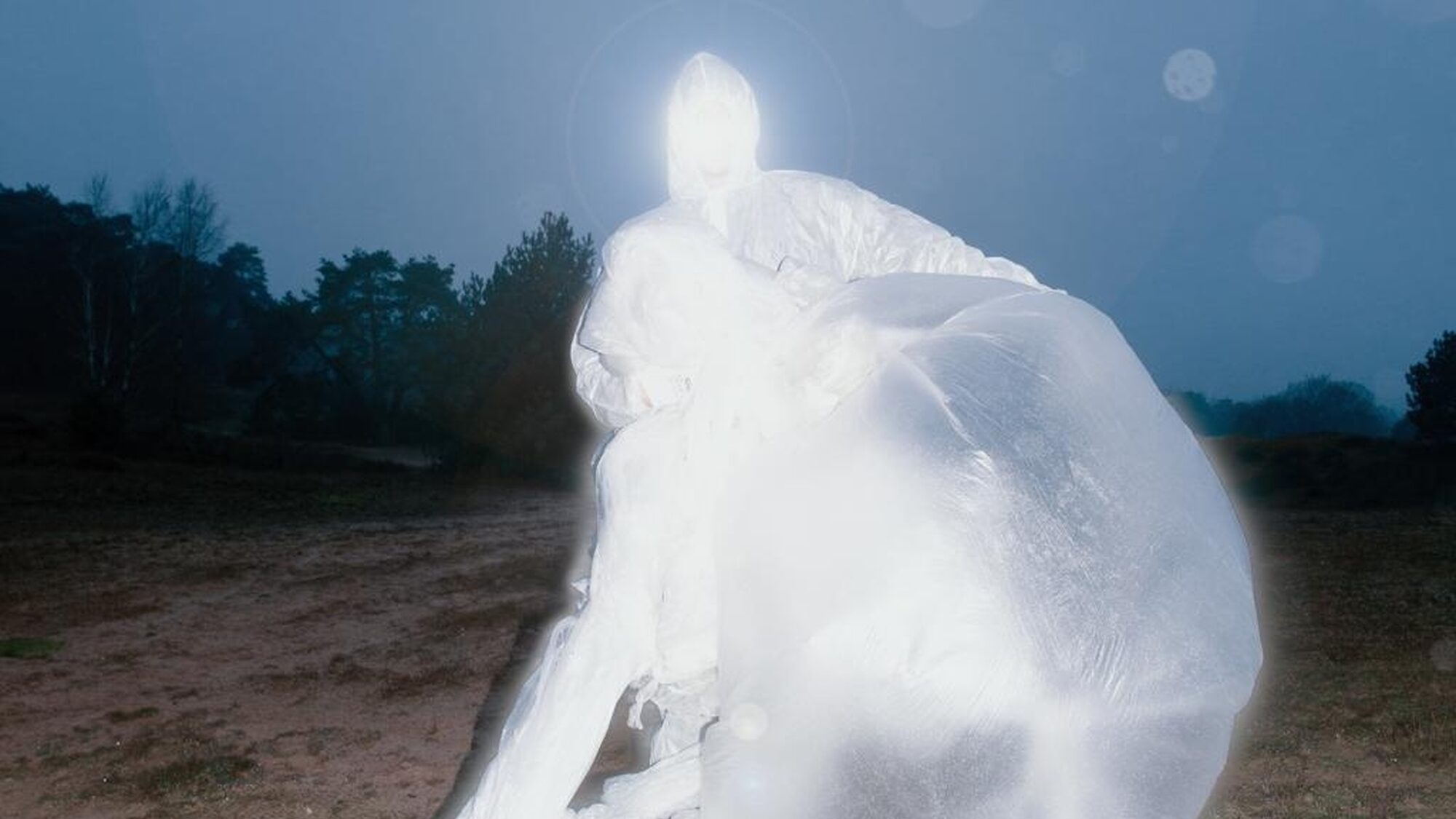 Een mens gehuld in een wit gewaad van materiaal dat op plastic lijkt. Je ziet niets van het gezicht. Op de achtergrond staan bomen.